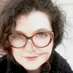 author headshot of a white woman with dark curly hair and tortoiseshell glasses. A lock of hair is across her face.