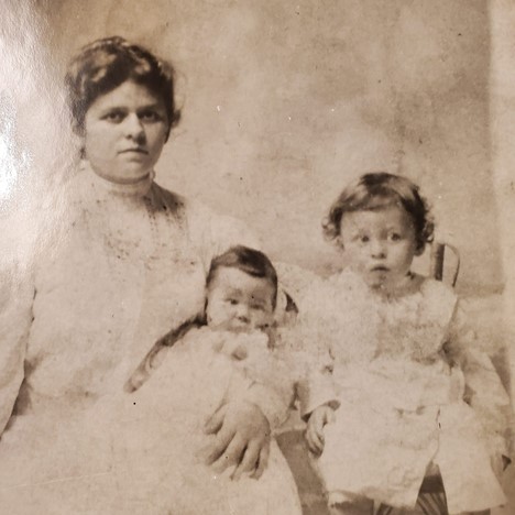 A sepia toned photograph of a dark haired woman holds an infant while a toddler sits beside them. All are wearing white frilly dresses.)